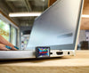 A small SANDISK Extreme Fit USB-C™ Flash Drive - 256 GB attached to the side of an open silver laptop on a wooden desk. A person's hand with teal nail polish is visible on the left, and the background shows a blurred indoor office or studio environment.