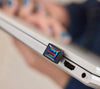 A close-up lifestyle shot of a SANDISK Extreme Fit USB-C™ Flash Drive - 256 GB attached to the side of a silver laptop, with a blurred hand holding the laptop in the background.