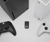 A SANDISK Optimus GX C50 Storage Expansion Card for XBOX Series X|S - 1 TB sits centered on a light gray surface, surrounded by parts of two Xbox consoles and two Xbox controllers in black and white.