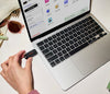 A hand holds a SANDISK USB-C Flash Drive - 256 GB  next to an open silver laptop on a white desk, with sunglasses and a bag partially visible in the background.