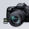 A black DSLR camera with a lens attached, shown on a light gray background with a small PNY EliteX-PRO60 UHS-II 512GB SDXC card displayed in front of it.